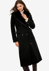 Abrigo de lana negro con cuello de piel sintética, doble botonadura en la parte delantera y cinturón en la cintura. Diseño de longitud media con mangas largas y bolsillos laterales.