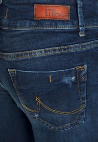 Jeans de denim azul escuro com etiqueta de couro LTB, costura amarela e design decorativo no bolso traseiro. Apresenta ligeiros detalhes desgastados.