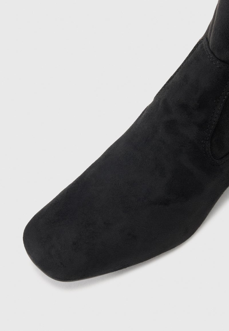 Czarne zamszowe botki sięgające kostki z okrągłym noskiem i gładką teksturą; minimalne wykończenia szwów; boczny elastyczny panel ułatwiający zakładanie.