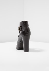 Musta nahkainen nilkkabootsi, jossa on korkea korko, sivuvetoketju ja koristeellinen solkiremmi. Liukas pinta ja sulavaliikkeinen muotoilu.