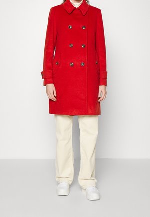 Osoba nosząca jasno czerwoną dwurzędową kurtkę, beżowe spodnie, białe buty, stojąca na jednolitym jasnym tle.