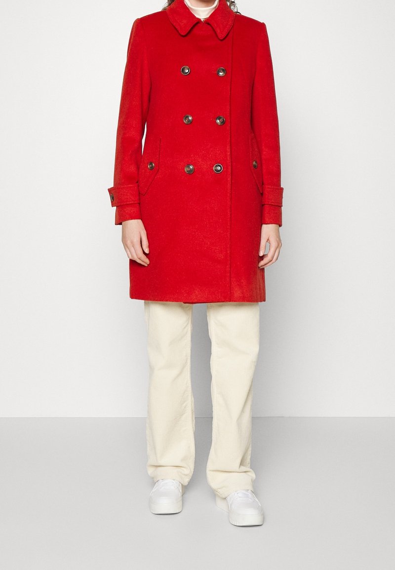 Personne portant un manteau croisé rouge vif, un pantalon beige, des chaussures blanches, debout devant un fond clair uni.