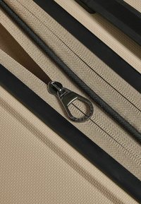 Exterior de equipaje beige con superficie texturizada, que cuenta con un zipper negro con una pestaña metálica y un ribete de tela negra en contraste.