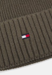 Olive green ribbed knit beanie featuring a small tri-color logo patch (red, white, blue) on the side. Soft texture, wide cuff.