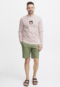 Felpa rosa chiaro con logo, pantaloni corti verdi e sandali marroni. La felpa ha polsini e vita a costine. Abbigliamento casual.