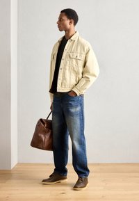 Hellblaues Jeansjackett über einem schwarzen Shirt, blaue Jeans und braune Schuhe. Eine braune Lederhandtasche wird an der Seite des Subjekts gehalten.