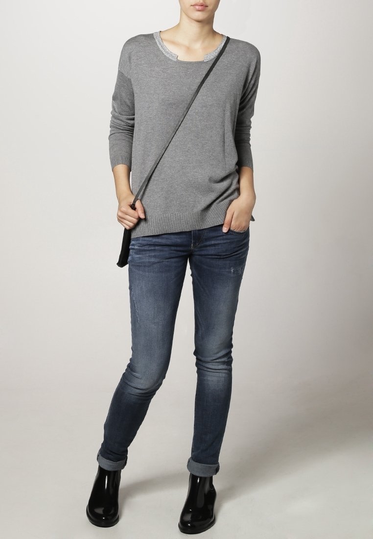 Grauer Strickpullover mit Rundhalsausschnitt und strukturiertem Saum. Kombiniert mit enganliegenden dunkelblauen Jeans und schwarzen Stiefeletten.