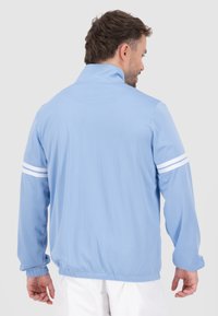 Giacca perforata azzurro chiaro con colletto alto, maniche lunghe e strisce bianche sul braccio sinistro. Polsini e orlo elasticizzati per un look aderente.