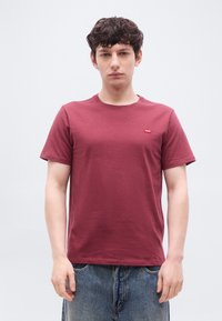 Ν έος άνδρας με σγουρά μαλλιά που φοράει απλό μαροον μπλουζάκι και μπλε τζιν παντελόνι, στέκεται μπροστά σε ένα απλό ανοιχτό γκρι φόντο.