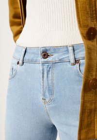 Ljusblå denimjeans med en figurnära design, som har en brun metallknapp och kontrasterande sömmar. Tillsammans med en krämfärgad ribbad topp och en senapsgul cardigan.