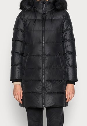 Czarna ocieplana zimowa kurtka z przednim zamkiem, dwoma zapinanymi na zamek kieszeniami oraz kapturem wykończonym futerkiem, noszona na czarnych spodniach.