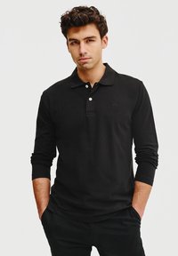Czarna koszulka polo z długim rękawem, uszyta z miękkiego materiału, z kołnierzykiem, trzema guzikami i subtelnym logo na klatce piersiowej. Prosty design.