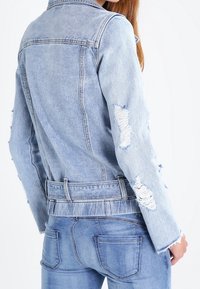 Žena v světle modré distressed denimové bundě a odpovídajících džínech, zobrazená zezadu na bílém pozadí.