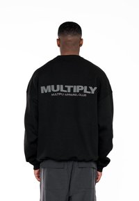 Černá mikina s velkým šedým nápisem na zádech, který zní "MULTIPLY" a "MULTIPLY APPAREL CLUB." Materiál vypadá měkce.