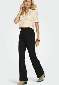 Blouse beige raccourcie avec deux poches poitrine et pantalons évasés taille haute noirs, associés à des bottines beige et des bracelets empilés.