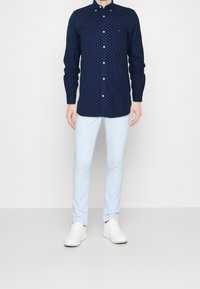 Tmavě modrá košile zapínaná na knoflíky s malým vzorem, kombinovaná se světle modrými kalhotami a bílými teniskami. Košile má límeček na knoflíky a jednu náprsní kapsu.