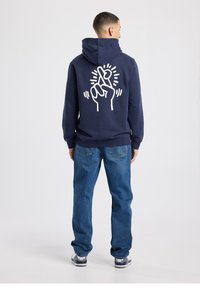 Navy hoodie in soft fabric features a graphic design of a hand crossing fingers on the back. Paired with loose blue jeans and sneakers.