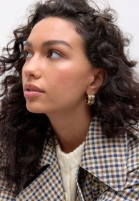 Gold hoop earrings with multiple rings, worn with a checkered beige and blue coat and a cream sweater, showcasing textured curly hair.