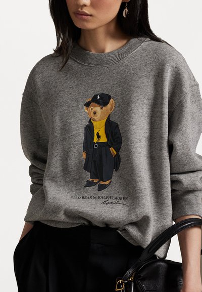 Pelēka sporta jakas ar attēlu, kurā redzams lācis dzeltenā rollē un melnā mēteli, ar tekstu "POLO BEAR by RALPH LAUREN" apakšā.