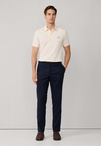 Cremfarbendes Poloshirt mit kurzen Ärmeln und Kragen, versehen mit einem kleinen blauen Logo. Kombiniert mit dunklen marineblauen gestreiften Hosen und braunen Slippern.