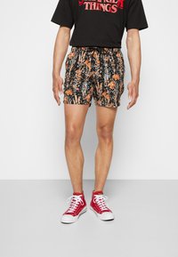 Homme portant un t-shirt noir de Stranger Things, un short noir à fleurs avec des fleurs orange et des baskets montantes rouges avec des lacets blancs.