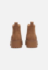 Bottines en cuir marron avec une doublure douce et texturée, semelle épaisse et plateforme, et une languette à l'arrière. Détail du logo sur le côté.