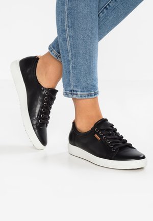 Pieds portant des baskets en cuir noir à lacets avec semelles blanches, associées à un jean bleu avec revers sur fond blanc.