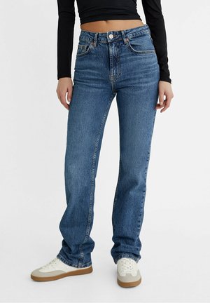Persoon draagt blauw, hoog getailleerde recht pantalon jeans, zwart cropped top met lange mouwen en witte sneakers met beige en bruine zolen.