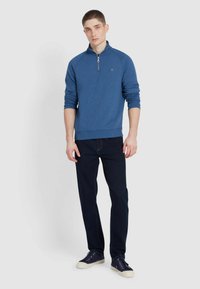 Blue zip-neck sweatshirt with ribbed cuffs and hem, paired with dark jeans and navy sneakers, featuring a small logo on the chest.