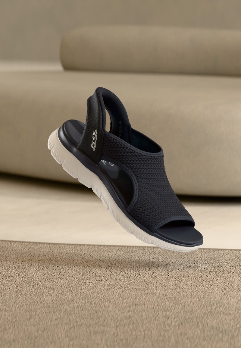 Μαύρο ανοιχτό δερμάτινο σανδάλι Skechers με υφασμάτινο επάνω μέρος με υφή και λευκή εύκαμπτη σόλα, εμφανίζεται σε φόντο μπεζ.