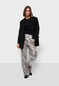 Zwarte blazer over een zwarte top, gecombineerd met glanzende zilveren wijde broek en zwarte schoenen. Gladde textuur en metallic afwerking op de broek.