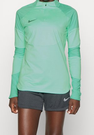 Person iført en atletisk langærmet top i teal med lynlås foran og sorte Nike-shorts med hvid swoosh-logo.