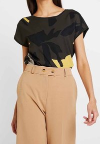 T-paita lyhyillä hihoilla tummanvihreänä, jossa on abstrakteja mustia ja keltaisia kuvioita; yhdistettynä vaaleanruskeisiin korkeavyötäröisiin housuihin, joissa on kaksi kultanappia.