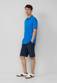 Blauwe poloshirt, marineblauwe shorts en witte sneakers met zwarte accenten. Korte mouwen, casual pasvorm, gladde stoftextuur.