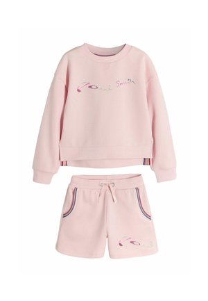 Young girl stands with hands in pockets wearing light pink sweatshirt and shorts, white socks, and sneakers against plain gray background.