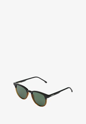 Lunettes de soleil en forme de chat avec une monture noire et des verres verts, présentant une accentuation en écaille de tortue. Branches noires fines. Design arrondi et anguleux.