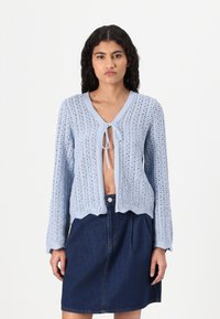 Cardigan lavorato a maglia azzurro chiaro con davanti aperto, chiusura a fiocco e bordi scallop, abbinato a una gonna in denim scuro.