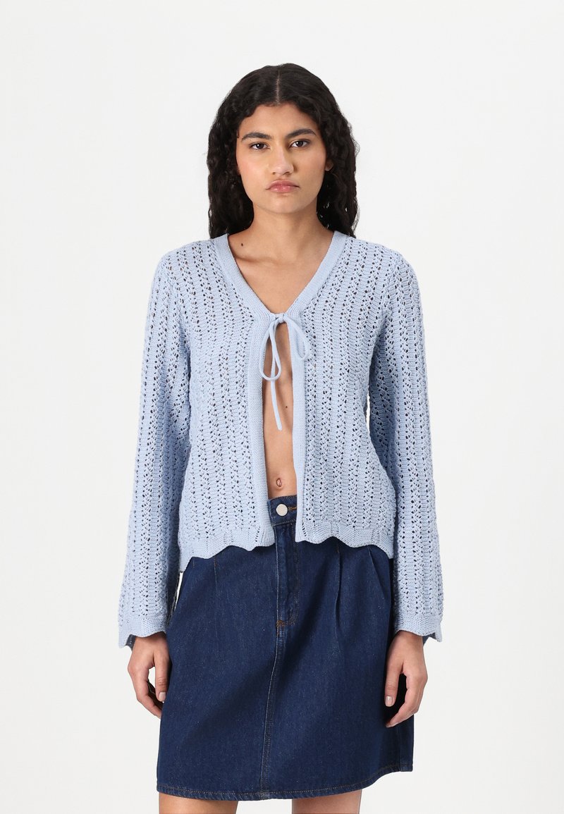 Cardigan lavorato a maglia azzurro chiaro con davanti aperto, chiusura a fiocco e bordi scallop, abbinato a una gonna in denim scuro.