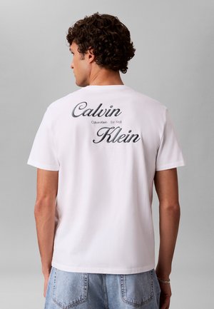 Mann mit lockigem Haar, der ein weißes Calvin Klein T-Shirt mit Logo auf dem Rücken trägt, kombiniert mit hellblauen Jeans, steht vor grauem Hintergrund.