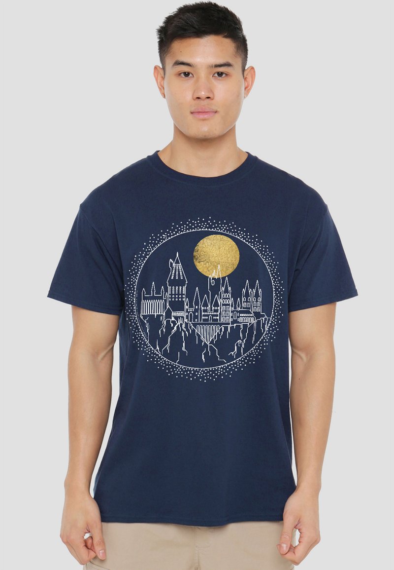 Man wearing navy blue t-shirt with white line art of a castle on cliffs and a large gold circle above it, central on the shirt.