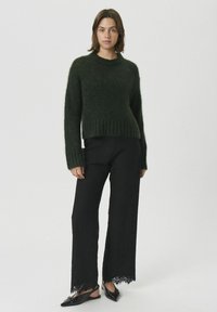 Dunkelgrüner, strukturierter Strickpullover mit langen Ärmeln und geripptem Saum. Kombiniert mit hoch taillierter schwarzer Hose mit Spitzenbesatz und spitzen schwarzen Schuhen.