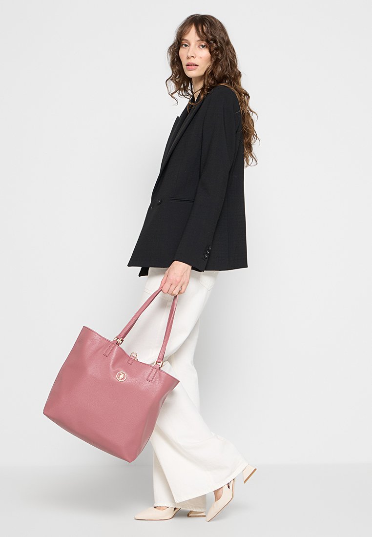 Bolso tote de cuero rosa con dos asas, acabado texturizado, detalles en dorado y logo. Combinado con una chaqueta negra y pantalones blancos.