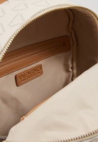 Intérieur d'un sac à dos en cuir beige avec doublure en tissu. Comprend une poche zippée, une étiquette de marque et une texture lisse. La fermeture éclair est de couleur dorée.