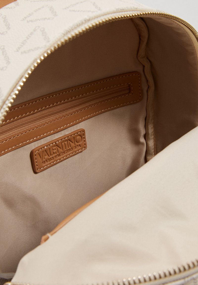 Interior de mochila de cuero beige con forro de tela. Cuenta con un bolsillo con cremallera, etiqueta de marca y textura suave. La cremallera es de tono dorado.
