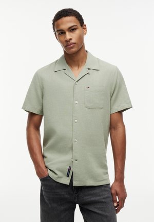 Άνδρας που φοράει ανοιχτό πράσινο πουκάμισο με κοντά μανίκια και τσέπη στο στήθος, στέκεται μπροστά σε απλό λευκό φόντο.