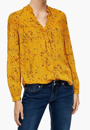 Kvinde iført gul langærmet bluse med flerfarvet abstrakt blomsterprint og blå jeans, stående med den ene hånd ved siden.