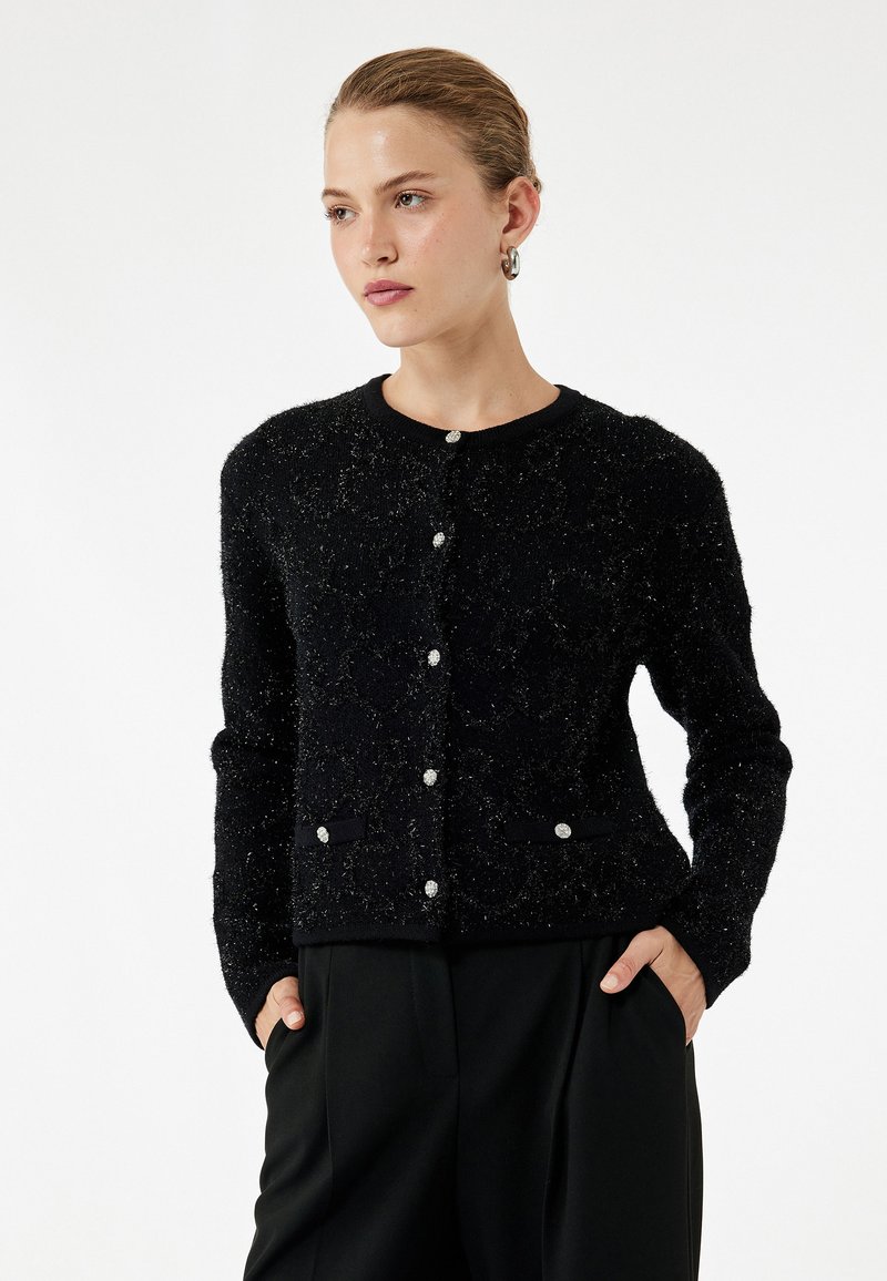 Schwarzer Cardigan aus strukturiertem Stoff mit einem subtilen Funkeln. Verfügt über einen runden Ausschnitt, vordere Knöpfe und Seitentaschen.