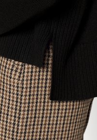 Svart ribbat stickat tröja överlappar bruna och svarta houndstooth-mönstrade byxor. Tröjan har en sidoklyvning nära fållen.