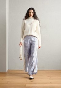 Un pull à col roulé côtelé crème, un pantalon large à motifs bleu clair, des ballerines noires, portant un sac blanc, se tenant sur un sol en bois.