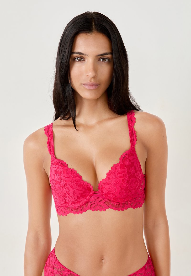 Bralette in pizzo di un vivace colore rosa, con bordi smerlati e motivi floreali, dotato di spalline regolabili e scollatura profonda.